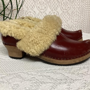 Pode incluir: Um par de tamancos com cabedal em couro bordô e sola de madeira clara. Os tamancos têm uma gola de pele de carneiro ao redor do tornozelo. Os sapatos estão em uma superfície branca estampada.