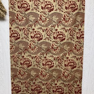May include: A beige fabric with a repeating pattern of red and brown stylized flowers. The flowers are arranged in a wavy pattern and are surrounded by smaller leaves and dots.