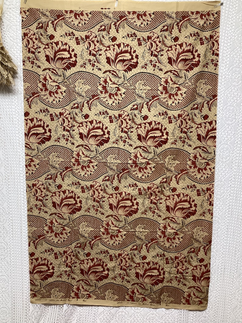 May include: A beige fabric with a repeating pattern of red and brown stylized flowers. The flowers are arranged in a wavy pattern and are surrounded by smaller leaves and dots.
