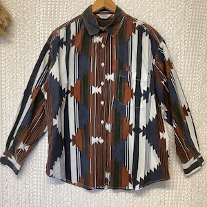 Peut inclure: Une chemise boutonnée à manches longues avec un motif géométrique dans les tons de marron, blanc, marine et vert. La chemise a un col et une poche poitrine unique. La chemise est suspendue à un cintre en bois.
