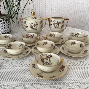 Puede incluir: Un juego de té vintage con fondo crema y detalles dorados. El juego incluye una tetera, una jarra de crema, un azucarero y seis tazas de té y platillos. Las tazas de té y los platillos presentan un diseño floral con pájaros.