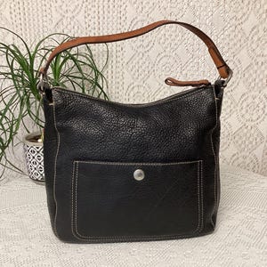Pu&ograve; includere: Borsa a tracolla in pelle nera con una texture martellata, una tasca frontale e un manico in pelle marrone. La borsa ha cuciture bianche e una chiusura con bottone color argento. Una pianta &egrave; visibile sullo sfondo.
