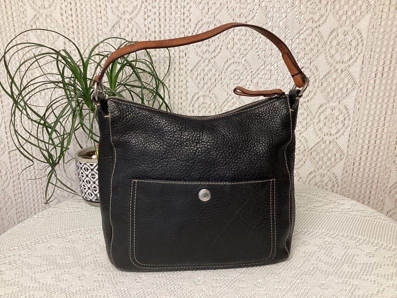 Pu&ograve; includere: Borsa a tracolla in pelle nera con una texture martellata, una tasca frontale e un manico in pelle marrone. La borsa ha cuciture bianche e una chiusura con bottone color argento. Una pianta &egrave; visibile sullo sfondo.