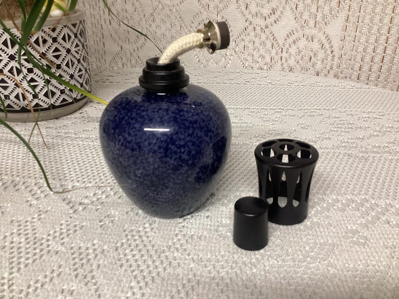 May include: A navy blue speckled ceramic oil lamp with a white wick and black accents. Includes a black metal cap and a small black cap. The lamp is designed for aromatherapy or decorative purposes.