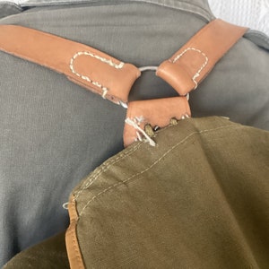 May include: A brown leather strap with white stitching is attached to a green canvas bag. The strap has a metal ring and is worn over the shoulders.