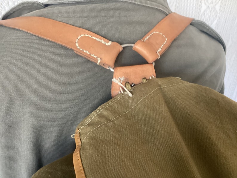 May include: A brown leather strap with white stitching is attached to a green canvas bag. The strap has a metal ring and is worn over the shoulders.