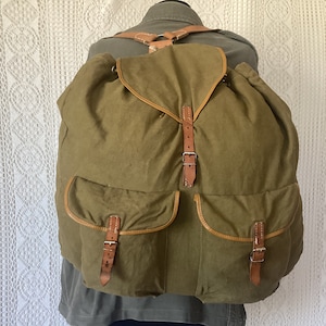 May include: A large, olive green canvas backpack with brown leather straps and buckles. The backpack has two large front pockets with brown leather trim.