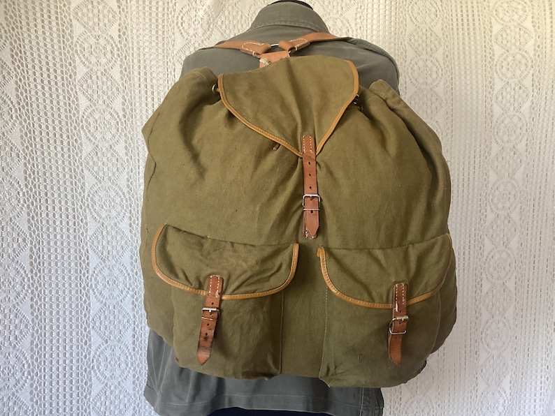 May include: A large, olive green canvas backpack with brown leather straps and buckles. The backpack has two large front pockets with brown leather trim.