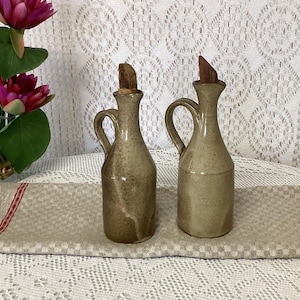 Peut inclure: Deux burettes en céramique avec un vernis beige moucheté, chacune dotée d'une anse incurvée, d'un col étroit et d'un bouchon en bois. Les burettes ont une base cylindrique et sont placées côte à côte, présentant une finition terreuse et lisse.