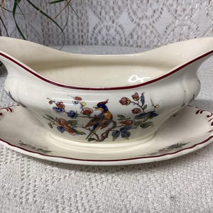 Op de afbeelding: Crèmekleurige juskom en bijpassende schotel met een bloemen- en vogelontwerp. De juskom heeft een gebogen vorm met een rode rand. De schotel is rond met een bijpassende rode rand en bloemmotief. Een decoratief serveerstuk.