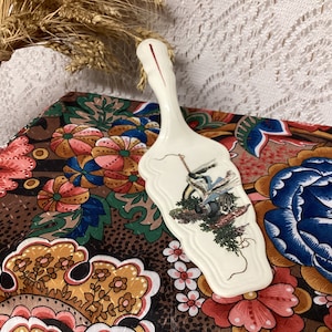 Puede incluir: Servidor de pastel de cerámica blanquecina con un mango decorativo y una escena pintada de un paisaje. El servidor se muestra sobre una tela estampada con diseños florales y de cachemira. Un manojo de trigo está en el fondo.