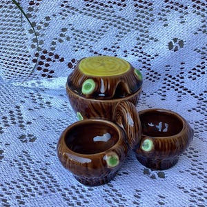 May include: A brown ceramic tea set with a tree stump design. The set includes a lidded teapot and two small cups. The teapot has a yellow top and green accents. The cups also have green accents.