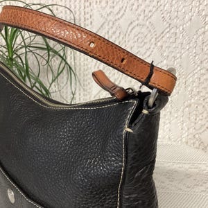Pu&ograve; includere: Borsa a tracolla in pelle nera con cinturino in pelle marrone. La borsa ha una superficie strutturata e cuciture bianche. Il cinturino &egrave; fissato con anelli in metallo argentato. Una piccola tasca &egrave; visibile sulla parte anteriore della borsa.