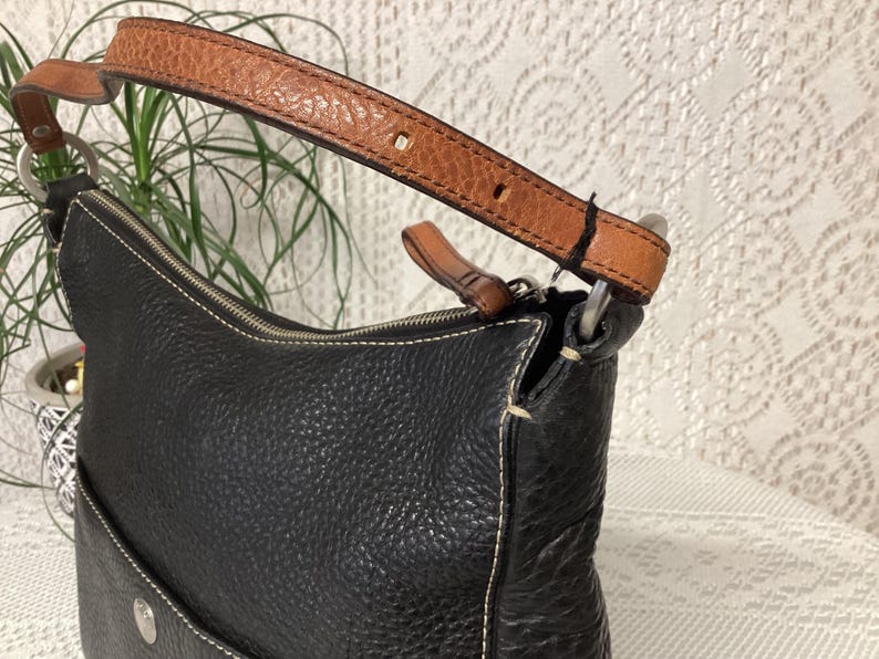 Pu&ograve; includere: Borsa a tracolla in pelle nera con cinturino in pelle marrone. La borsa ha una superficie strutturata e cuciture bianche. Il cinturino &egrave; fissato con anelli in metallo argentato. Una piccola tasca &egrave; visibile sulla parte anteriore della borsa.