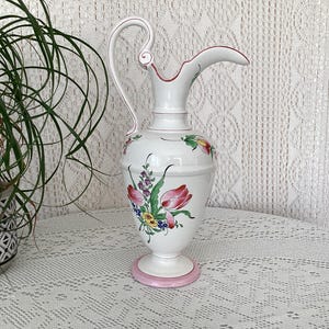 May include: A white ceramic pitcher with a pink base and floral designs in shades of pink, red, yellow, and green.