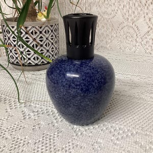 May include: A blue speckled ceramic fragrance lamp with a black top. The lamp has a rounded body and a decorative black top. The lamp is sitting on a white lace tablecloth. A potted plant is in the background.