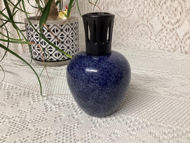 May include: A blue speckled ceramic fragrance lamp with a black top. The lamp has a rounded body and a decorative black top. The lamp is sitting on a white lace tablecloth. A potted plant is in the background.