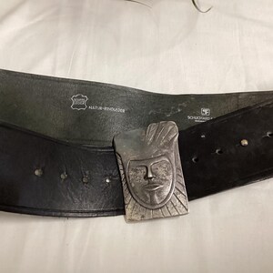 May include: A black leather belt with a silver-colored buckle featuring a stylized face design. The belt has several holes for adjustment and is marked with "Echte Leder" and "Natur-Rindleder".