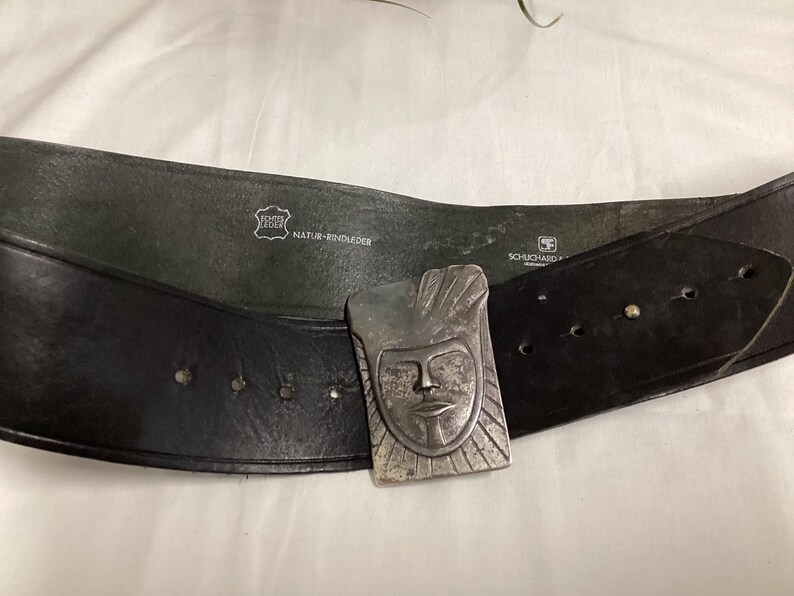 May include: A black leather belt with a silver-colored buckle featuring a stylized face design. The belt has several holes for adjustment and is marked with "Echte Leder" and "Natur-Rindleder".