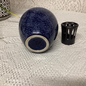 May include: A navy blue speckled ceramic vase with a white rim, next to a black metal candle holder. The vase has a rounded shape and a speckled pattern. The candle holder has a cylindrical shape with cut-out designs.