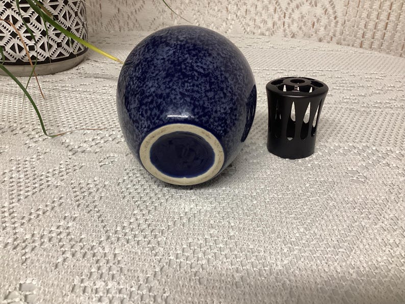 May include: A navy blue speckled ceramic vase with a white rim, next to a black metal candle holder. The vase has a rounded shape and a speckled pattern. The candle holder has a cylindrical shape with cut-out designs.