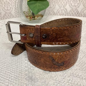 Pode incluir: Um cinto de couro castanho com fivela prateada. O cinto apresenta desenhos florais e de rolagem em relevo, com as palavras "EL CHARRO" na frente. O cinto tem aproximadamente 3,8 cm de largura.