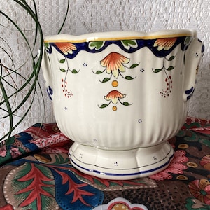 Może przedstawiać: Biała ceramiczna doniczka z niebieskimi, zielonymi, pomarańczowymi i żółtymi wzorami kwiatowymi. Doniczka ma dwa uchwyty i falisty brzeg.