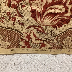 May include: A close-up of a fabric with a floral pattern in shades of brown and beige. The fabric has a repeating pattern of stylized flowers and leaves. The fabric is labeled "ANDUZE" and "un imprime par Roland Saget" in French.
