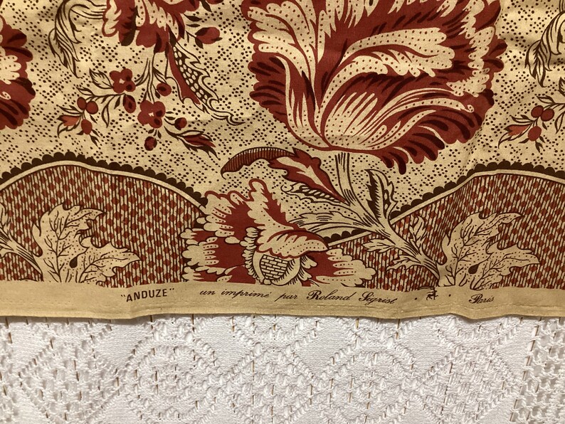 May include: A close-up of a fabric with a floral pattern in shades of brown and beige. The fabric has a repeating pattern of stylized flowers and leaves. The fabric is labeled "ANDUZE" and "un imprime par Roland Saget" in French.