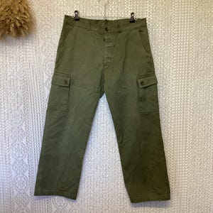 Puede incluir: Pantalones cargo de color verde oliva con cierre de botones, dos bolsillos laterales tipo cargo con solapas abotonadas y dos bolsillos laterales estándar. Los pantalones tienen una cintura estructurada con trabillas y cuelgan de una percha.