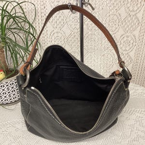 Pu&ograve; includere: Borsa a tracolla in pelle nera con cinturino in pelle marrone e finiture argentate. La borsa ha una chiusura a zip e cuciture bianche. L'interno &egrave; nero. Una pianta in un vaso decorativo &egrave; sullo sfondo.