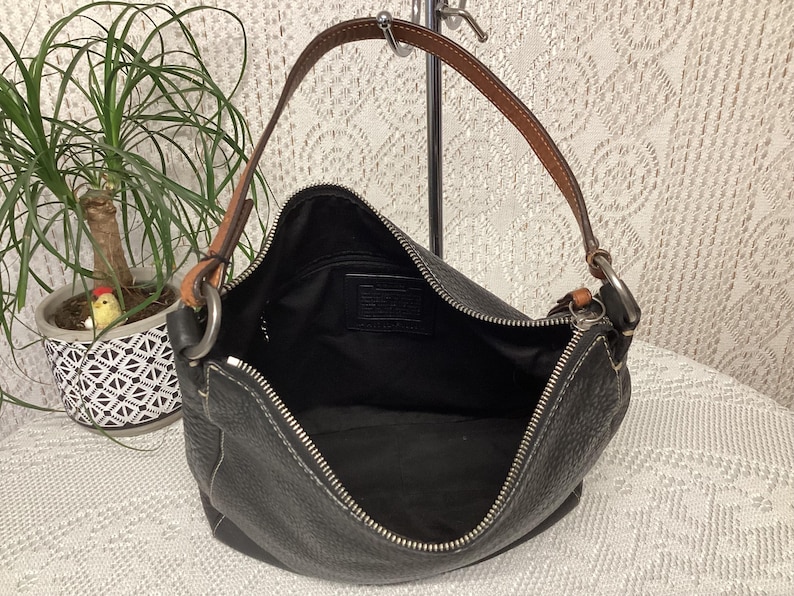 Pu&ograve; includere: Borsa a tracolla in pelle nera con cinturino in pelle marrone e finiture argentate. La borsa ha una chiusura a zip e cuciture bianche. L'interno &egrave; nero. Una pianta in un vaso decorativo &egrave; sullo sfondo.