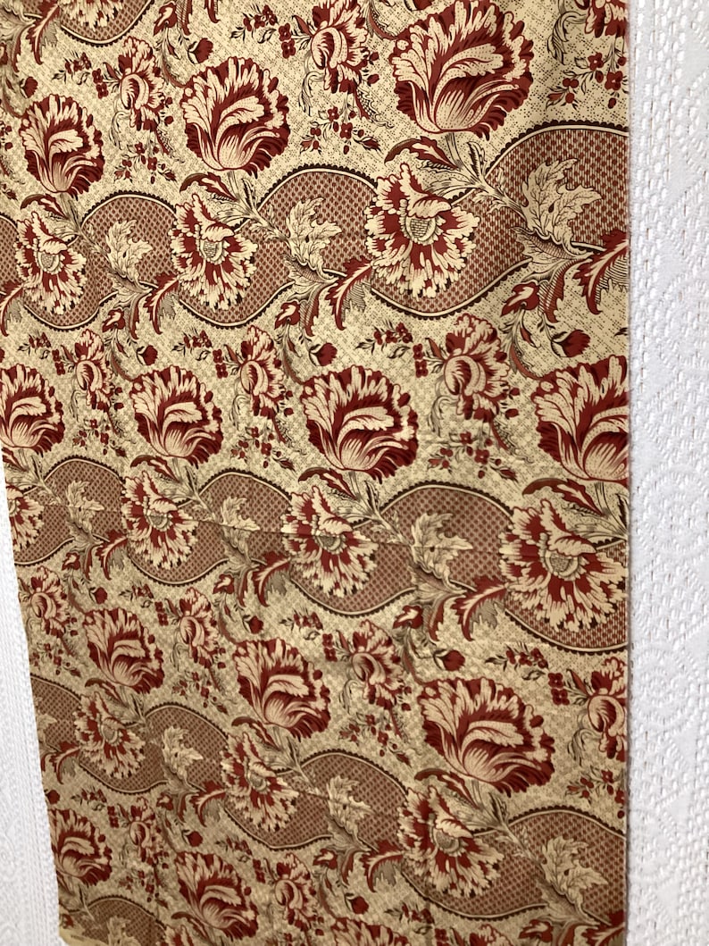 May include: A beige fabric with a repeating pattern of red and beige stylized flowers. The flowers are arranged in a wavy pattern.