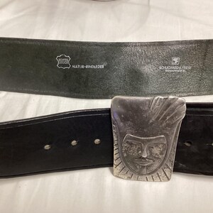 May include: Black leather belt with a silver-colored buckle featuring a stylized face design. The belt has a textured appearance and visible stitching. The back of the belt is dark green with text that reads "Echte Leder" and "Natur-Rindleder".