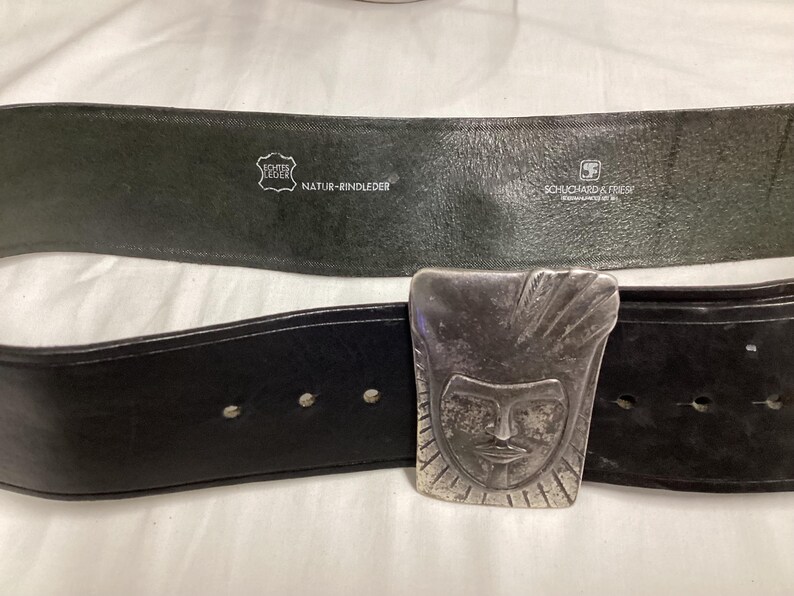 May include: Black leather belt with a silver-colored buckle featuring a stylized face design. The belt has a textured appearance and visible stitching. The back of the belt is dark green with text that reads "Echte Leder" and "Natur-Rindleder".