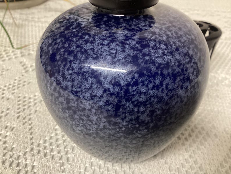 May include: A dark blue ceramic vase with a speckled white pattern. The vase has a rounded shape and a black top. The surface is glossy and reflects light. The vase is likely for home decor.