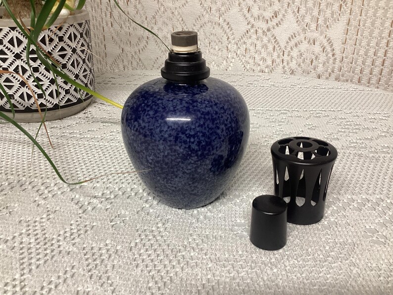 May include: A navy blue speckled ceramic oil lamp with a black top and a black metal decorative sleeve. A small black cap sits next to the lamp. The lamp is a decorative home decor item.