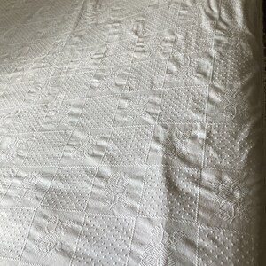 May include: White, textured quilt with a repeating pattern of squares and floral designs. The quilt has a raised, embossed appearance, with a variety of textures within each square. The overall design is symmetrical and classic, suitable for a bedspread or decorative throw.