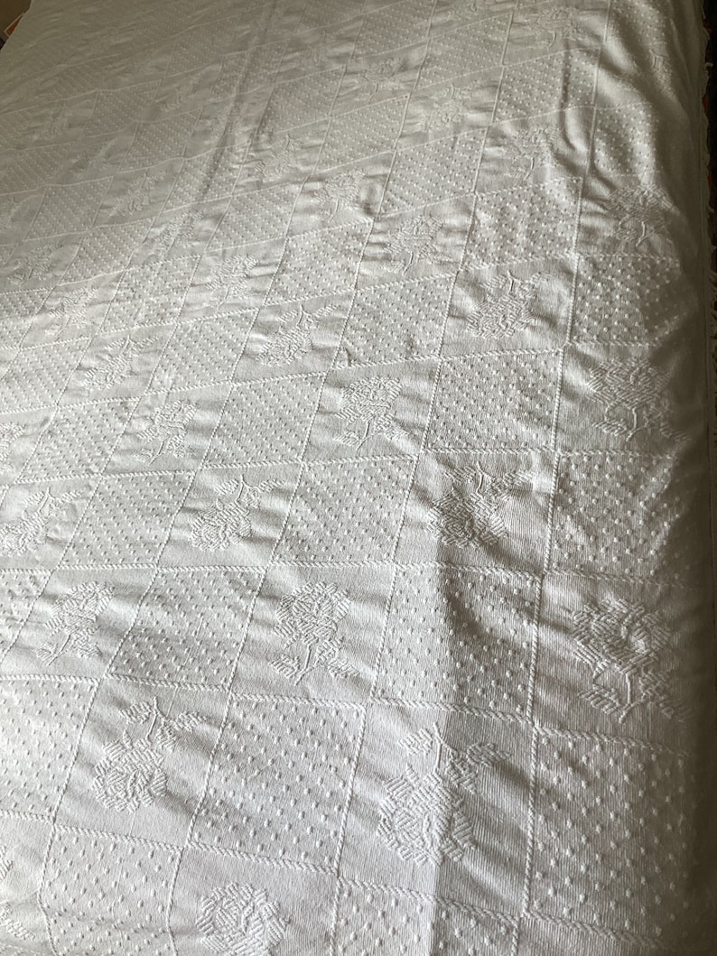 May include: White, textured quilt with a repeating pattern of squares and floral designs. The quilt has a raised, embossed appearance, with a variety of textures within each square. The overall design is symmetrical and classic, suitable for a bedspread or decorative throw.