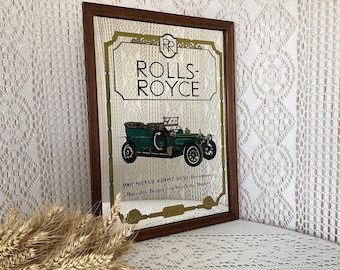 May include: A framed mirror featuring a vintage Rolls-Royce advertisement. The image showcases a green classic car with the text "ROLLS-ROYCE" at the top. The frame is dark brown, and the background is a cream color.