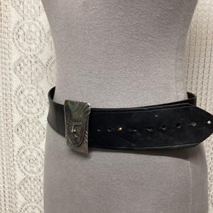 May include: Black leather belt with a silver-toned buckle featuring a stylized face design. The belt is worn around a mannequin. The buckle is rectangular and has a detailed, embossed pattern. The belt has several holes for adjustment.
