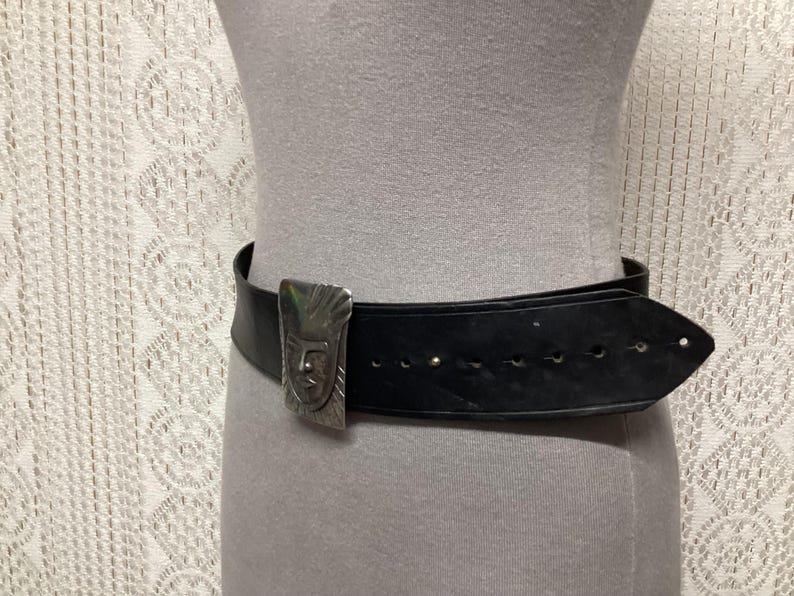 May include: Black leather belt with a silver-toned buckle featuring a stylized face design. The belt is worn around a mannequin. The buckle is rectangular and has a detailed, embossed pattern. The belt has several holes for adjustment.
