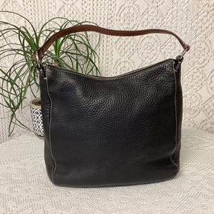Pu&ograve; includere: Borsa a tracolla in pelle nera con manico marrone. La borsa ha una superficie strutturata e cuciture bianche. La borsa &egrave; su una tovaglia di pizzo bianco.