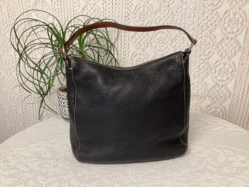 Pu&ograve; includere: Borsa a tracolla in pelle nera con manico marrone. La borsa ha una superficie strutturata e cuciture bianche. La borsa &egrave; su una tovaglia di pizzo bianco.