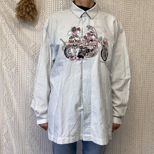 May include: A light blue long-sleeved button-down shirt with a cartoon graphic of two characters riding a motorcycle. The characters are wearing helmets and goggles.