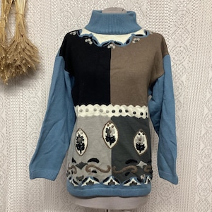 Puede incluir: Un suéter vintage con diseño de patchwork. El suéter presenta un cuello alto y mangas azules. El panel frontal está dividido en secciones negras, marrones, grises y blancas, con patrones de hojas y flores.