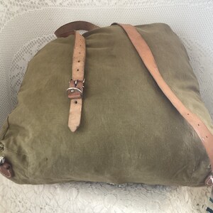 May include: A green canvas backpack with brown leather straps and a silver buckle. The backpack is lying on a white lace surface.