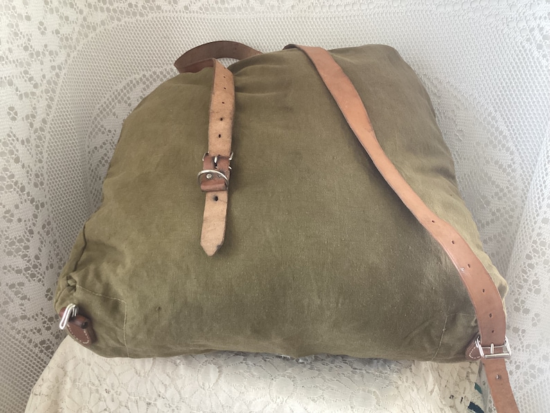 May include: A green canvas backpack with brown leather straps and a silver buckle. The backpack is lying on a white lace surface.