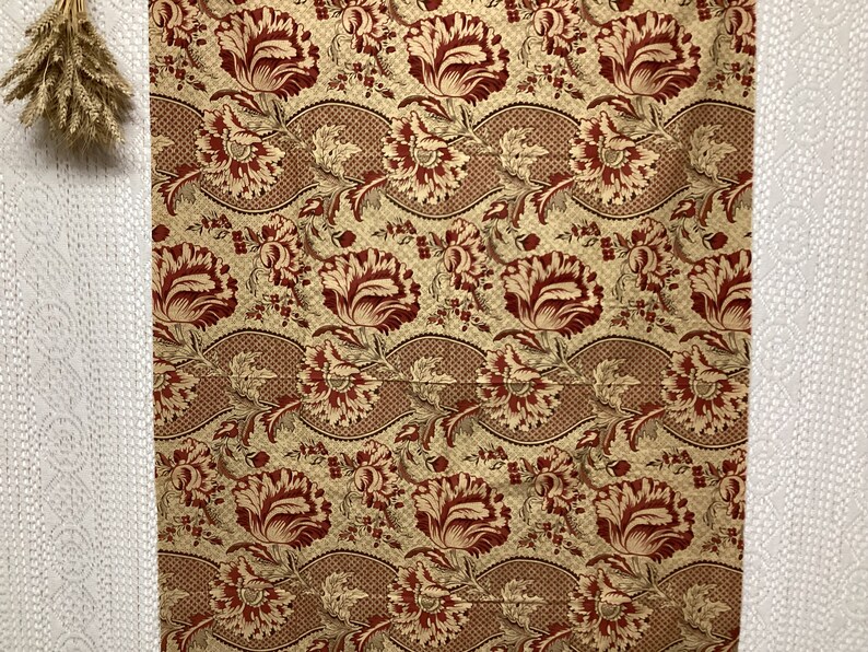 May include: A fabric swatch with a repeating pattern of red and cream floral designs on a beige background.