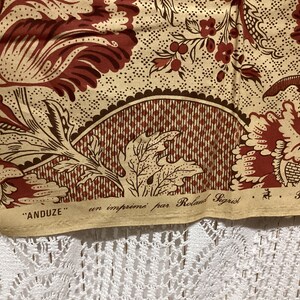 May include: Close-up of a beige fabric with a brown and red floral pattern. The fabric has a label that reads "ANDUZE" en imprim&eacute; par Roland Sigrist Paris.